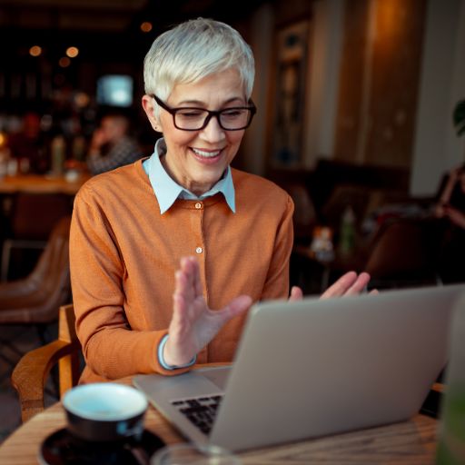 A woman wtih grey hair and glasses sitting at a cafe using her laptop and smiling