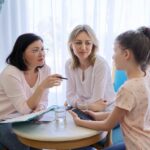 Female practitioner, talking to mother and daughter clients at a small round table. A glass of water sits in middle of table. The mother is smiling and looking at her daughter.