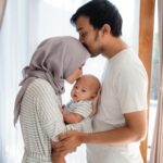 Parents hold their baby together. The mother holds her baby and kisses it on the forehead. The father rests his hands on the mothers arms, and kisses her forehead.