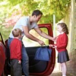 father standing near drivers door of a red car, handing lunch box to daughter in red school jumper. Son stands near father also in red school jumper.